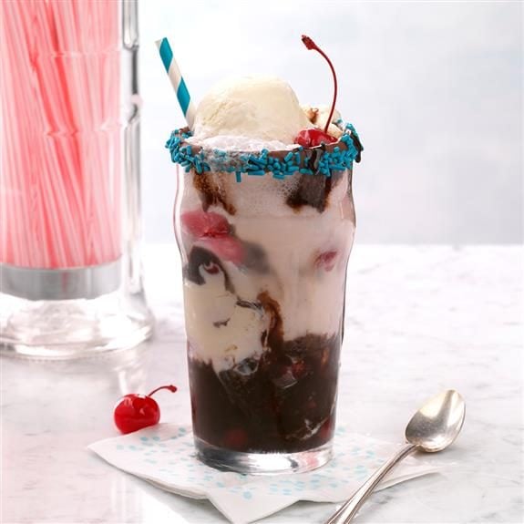 Float with swirls of chocolate syrup and cherries inside. The top of the glass is rimmed with chocolate and blue sprinkles and a matching striped straw rests inside