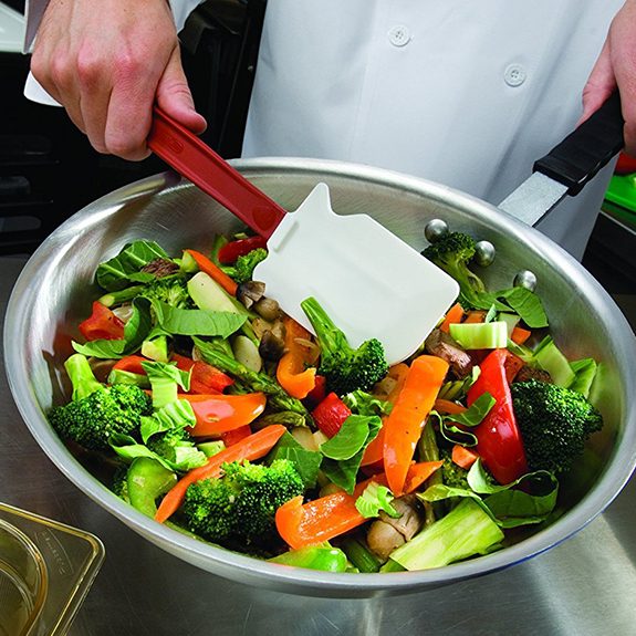 Red spatula being used to stir a skillet full of sliced veggies over a metal counter