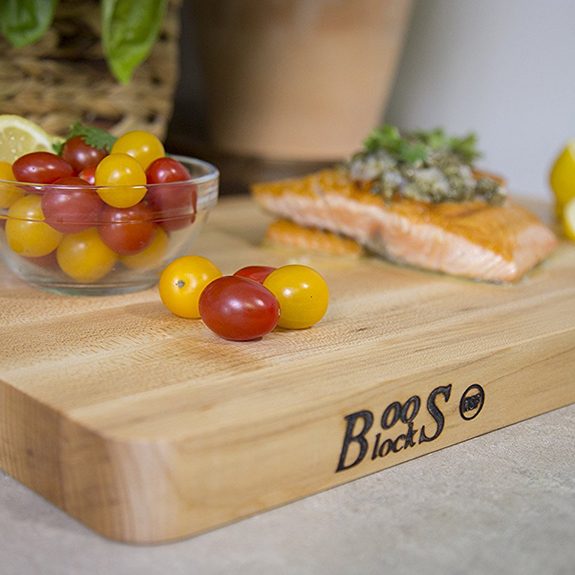 Thick, wooden cutting board with the word 'Boos Block' burned into the left corner and cherry tomatoes in a glass bowl and salmon resting on top of it