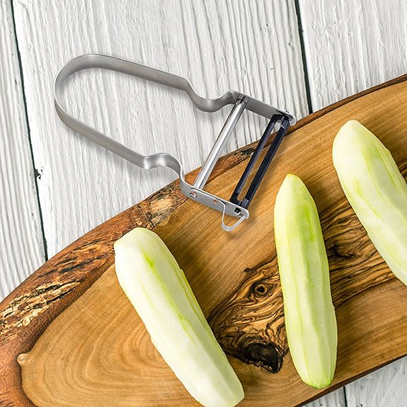 Vegetable peeler half on a wooden cutting board with three peeled zucchini lined up