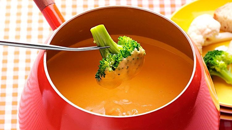 Person dipping broccoli into a red pot of cheese fondue
