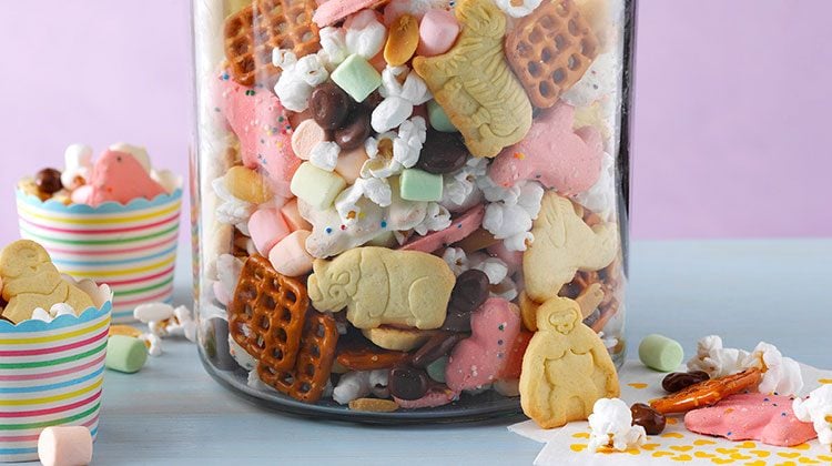 Glass jar filled with animal crackers, pretzels, popcorn, and other candies. The mixture is also loose around the base of the jar and in a couple striped paper cups