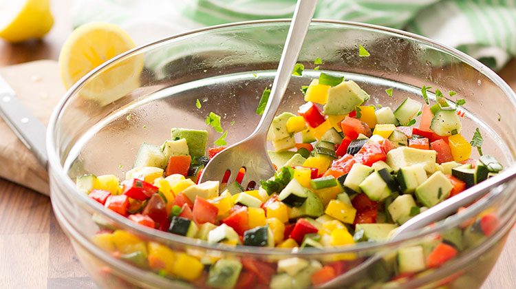 Chop salad made up of cubed cucumbers, tomatoes, avocado and peppers sitting in a glass bowl with two serving spoons