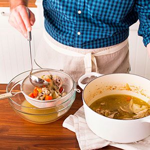 Person pouring the finished broth through a strainer over top a glass bowl using a large spoon