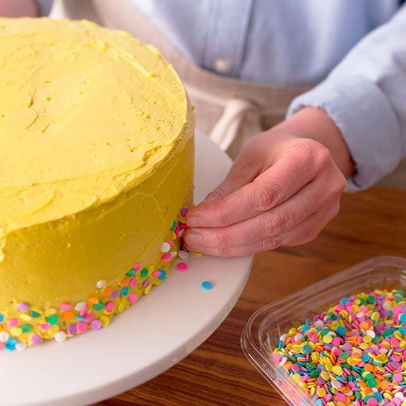 funfetti cake recipe, colorful sprinkles are being stuck onto the yellow buttercream frosting of a cake