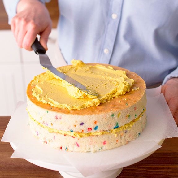 funfetti cake recipe, yellow buttercream frosting is being spread on top a sprinkle-filled cake
