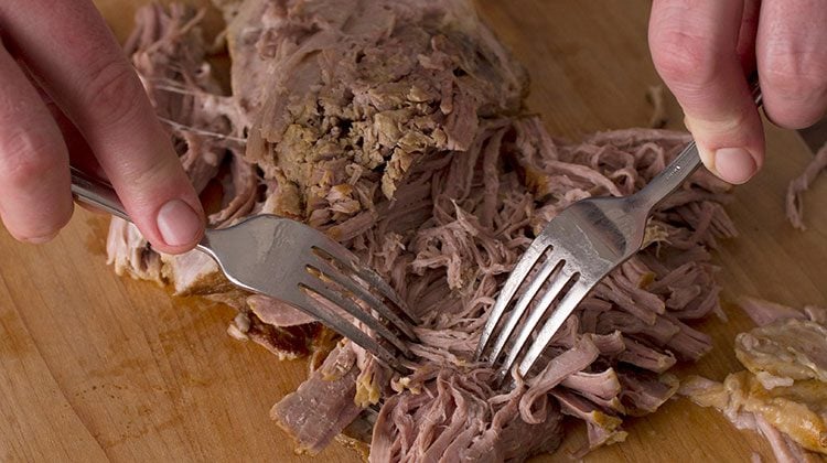man using two forks to carefully shred a portion of a pork shoulder