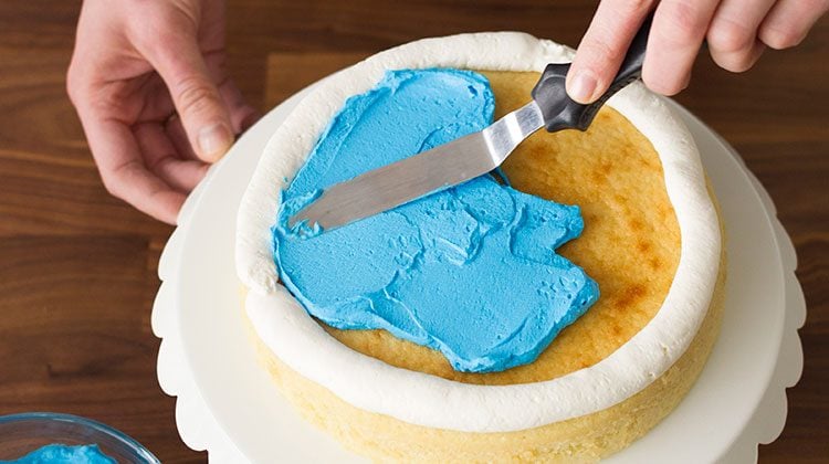 Person spreading a cup of blue frosting into the middle of a thick layer of white frosting