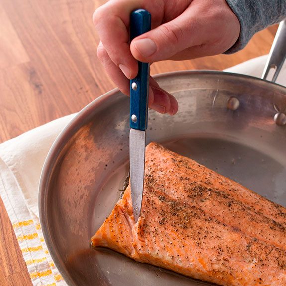 Filet of salmon cooking in a skillet as a person inserts a knife to check if the filet flakes