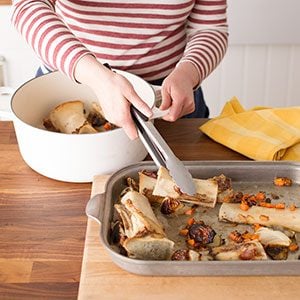 Using metal tongs, a person moves the bones from the baking sheet back into the stockpot