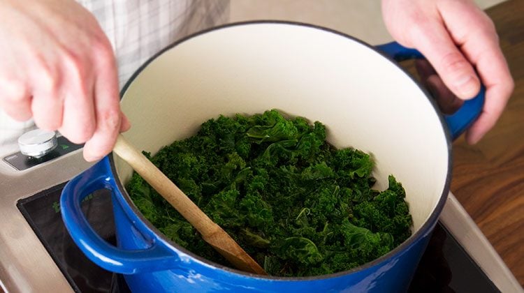 kale simmering in a large blue pot on a stove top as a person stirs it