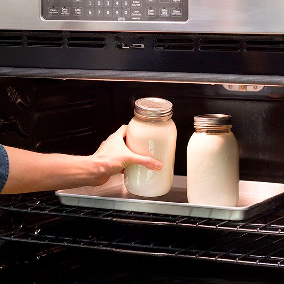 Two large mason jars filled with yogurt being placed into a metal pan in an oven