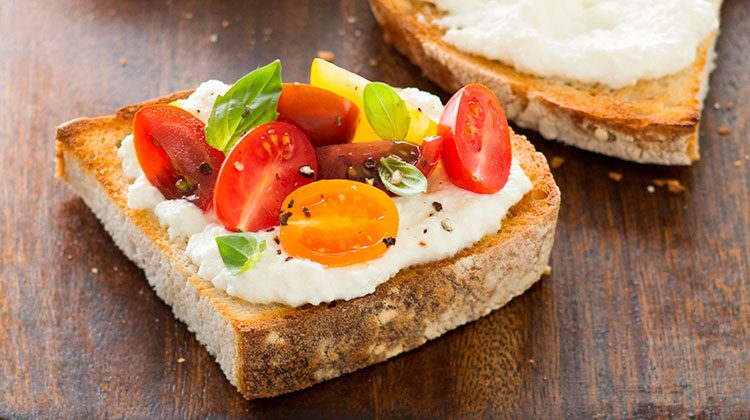 Piece of bread with ricotta spread liberally over it and topped with halved cherry tomatoes