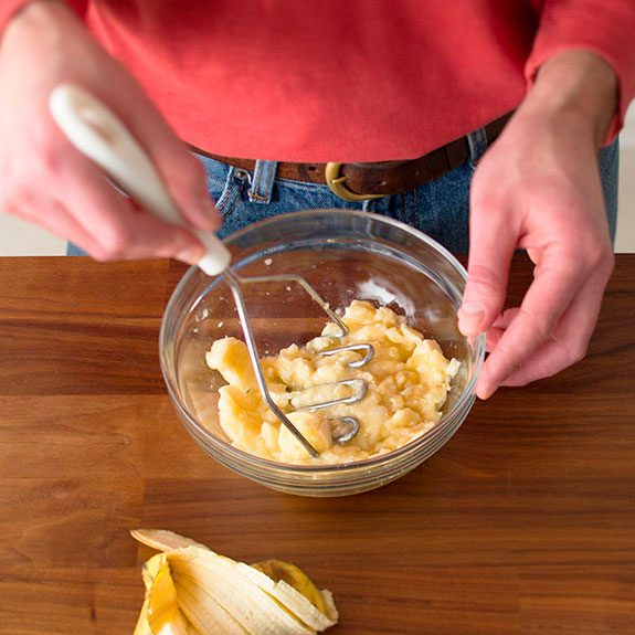 person using a potato masher to mash bananas in a glass bowl