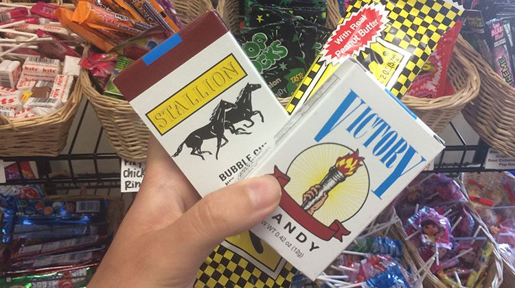 Hand holding a Victory brand and Stallion brand candy cigarettes against a backdrop of baskets filled with other old-fashioned candies on shelves