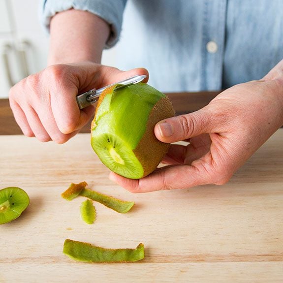 person holding a kiwi and peeling off its skin with a vegetable peeler
