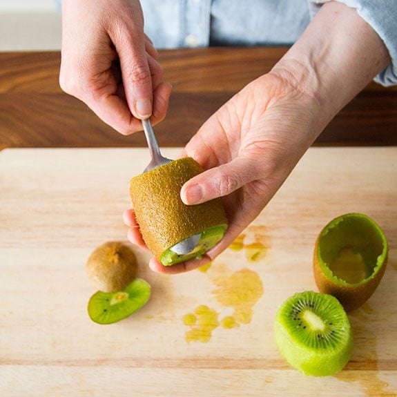 person holding a kiwi and using a spoon to get underneath its skin