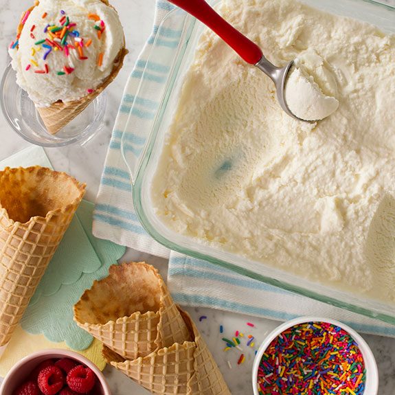 scattered assortment of waffle cones, sprinkles and raspberries beside a pan of ice cream