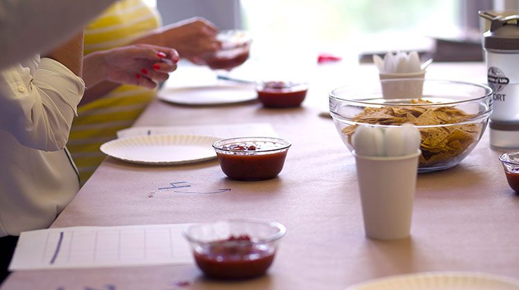 People standing around a table with a row of plastic bowls filled with salsa and one large glass bowl of tortilla chips