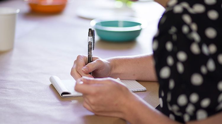 Person writing on a pad of paper with a pen with a blue bowl in the background