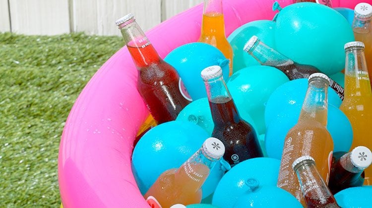 Soda bottles in a pink and yellow stripped, blow-up kiddie pool along with blue water balloons to keep them cool