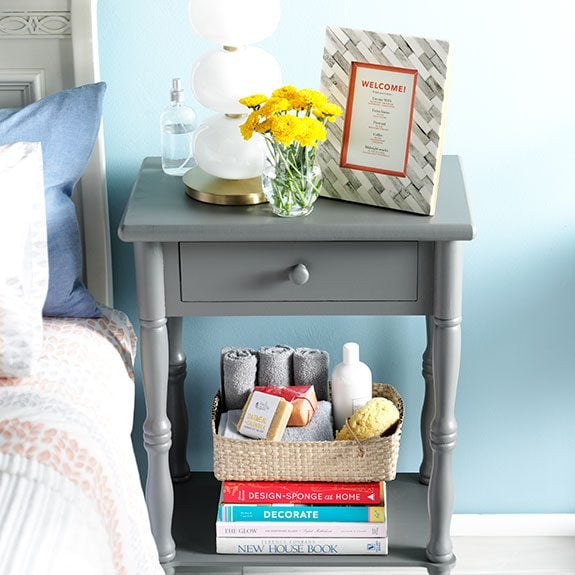 Night stand with a framed welcome note and flowers on top and books with assorted tolietries on a lower shelf