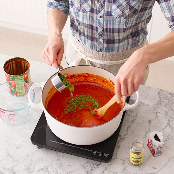 Person sprinkling in herbs into a pot on the stovetop with one hand and stirring sauce with a wooden spoon with their other