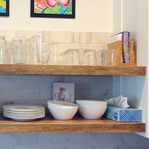 Two layers of wooden shelving installed on a kitchen wall and lined carefully with stacked glasses, bowls, and plates