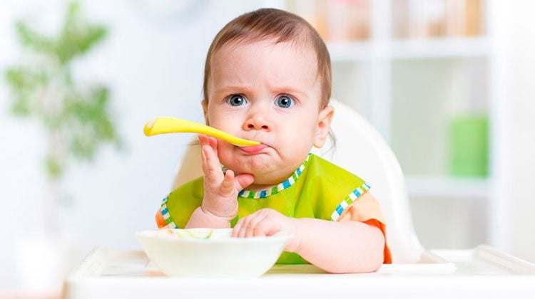 Frowning baby with a yellow spoon sticking out of their mouth. One hand is reaching up to hold their chin and the other is reaching into their white bowl on the tray of their high chair