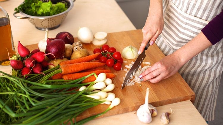 Person dicing onions with a kitchen knife on a wooden cutting board with other vegetables including radishes, potatoes, red onions, and carrots wait on the side