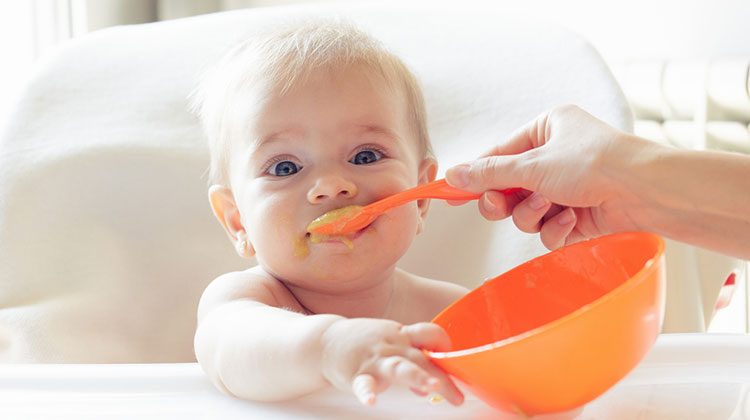 Baby look straight at the camera, one hand reached out to the edge of an orange bowl in front of them while a spoon filled with yellow mush is being pushed into their mouth