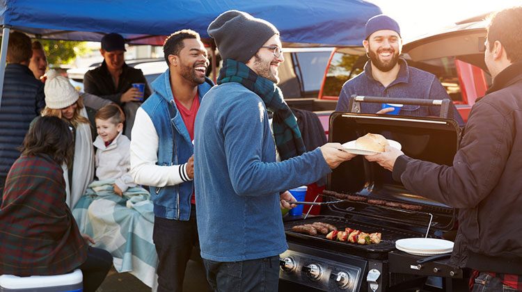 People dressed in sweaters, coats and laughing gathered around a grill filled with sausages