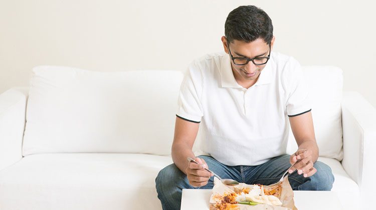 Man sitting alone on a white sofa and digging into a plate of food in front of him on a coffee table
