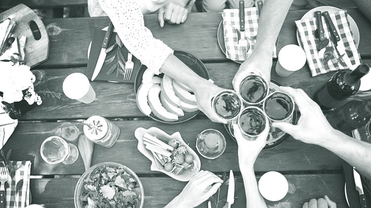 Black and white picture of a wooden picnic table covered with plates of food including slices of fruit, salads and bottles of wine. Four humans clink their glasses together