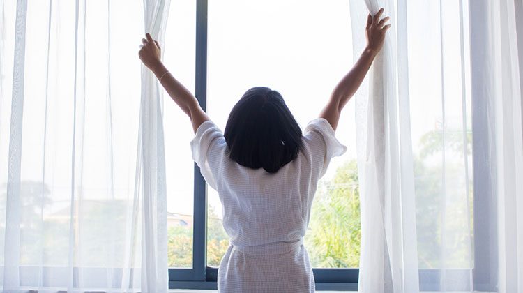 Woman with shoulder length hair and wearing a white robe reaching up to pull back curtains to let in the morning light