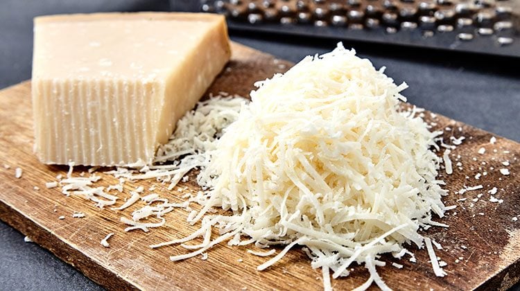 Chunk of cheese on a wooden cutting board beside a pile of shreds with a grater behind them