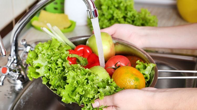 Hands holding a metal strainer full of red peppers, onions, lettuce leaves, tomatoes and oranges under a running faucet