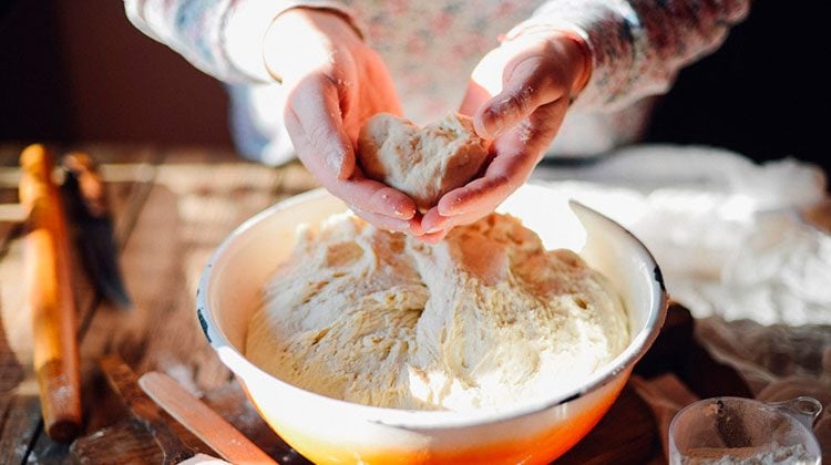 Bare hands reaching in and pulling out a heart-shaped lump of dough from the main dough from a large orange bowl