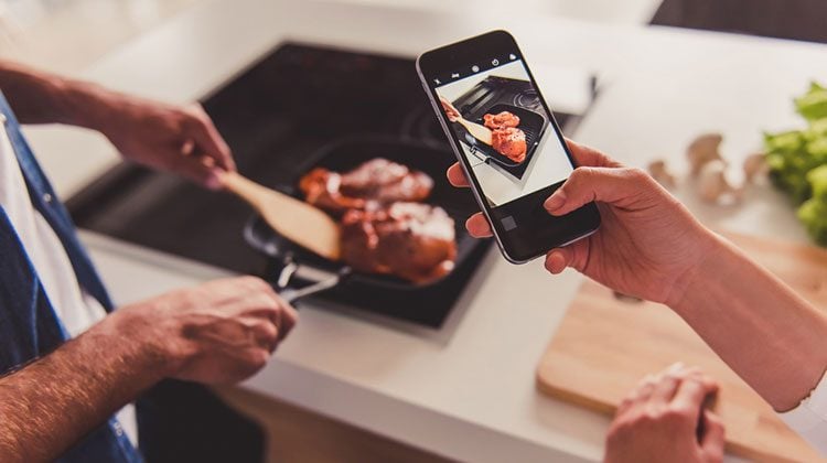 One person holding up a smartphone and filming another person as they grill meat in a skillet on the stovetop