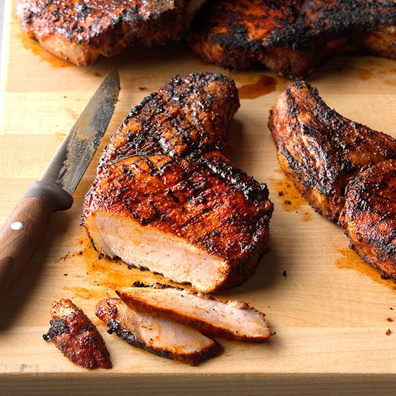 Two perfectly grilled pork chops on wooden cutting board. The pork chop on the left has several slices cut into its top