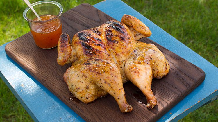 A grilled spatchcock chicken on a wood board upon a small blue table with a dressing in a glass jar and spoon off to the side