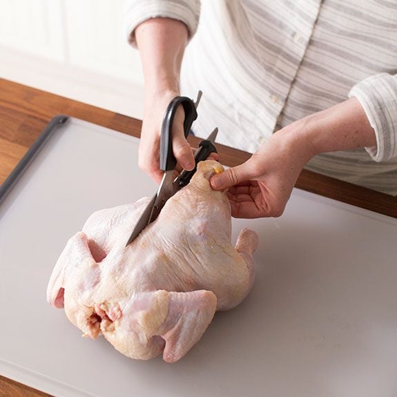 Raw chicken belly down on a white cutting board being opened with a pair of kitchen shears