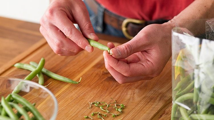 Person carefully removing the withered ends from a green bean with their hands. The ends are gathered on a cutting board below them and the finished beans are in a glass bowl