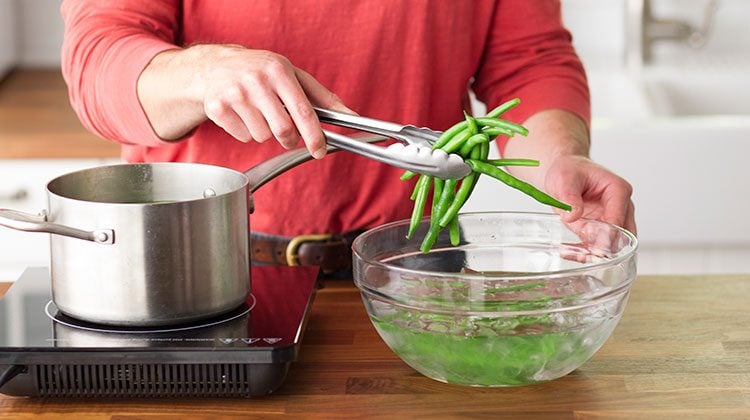 Person using metal tongs to remove green beans from a cold water bath in a glass bowl to place them in a metal pot on the stovetop