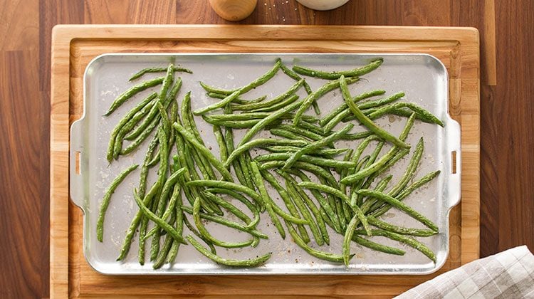 Green beans spread out over a baking sheet resting on a wooden cutting board with a gray checkered towel on its lower right corner