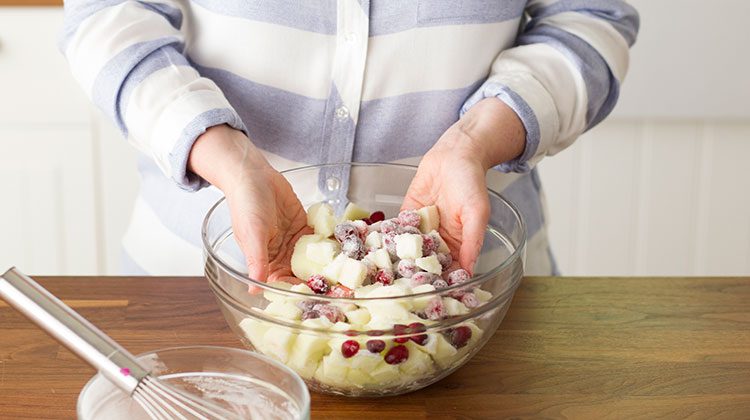 Hands mixing different fruits together in a glass bowl with a sugar mixture to coat them