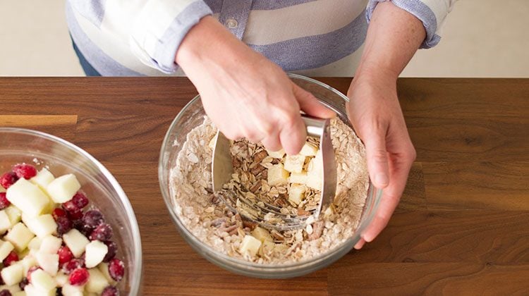 Hands using a tool to mash cubed butter into oats, brown sugar and nuts in a glass bowl