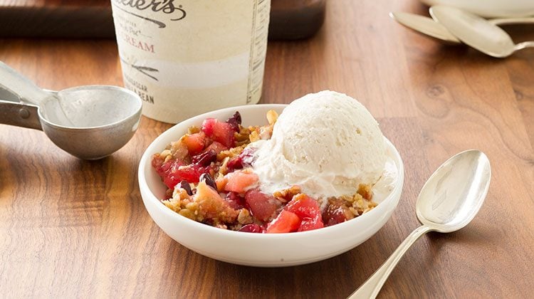 Fruit crisp beside a generous scoop of vanilla ice cream in a white bowl with a spoon ready at its side