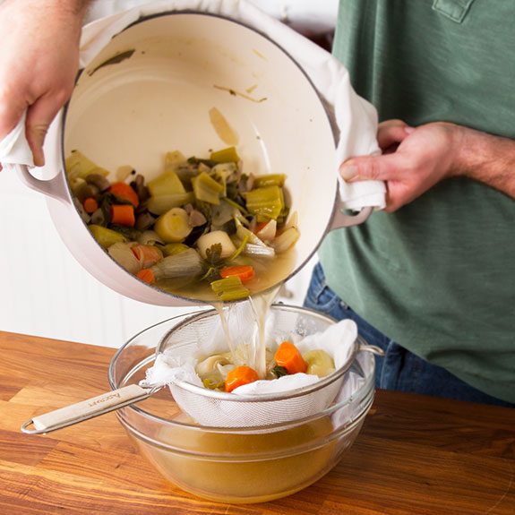 Person pouring the broth from the stock pot into a colander lined with cheesecloth to catch the vegetables