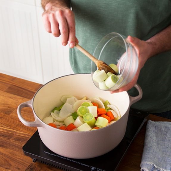 Person pushing sliced vegetables out of a glass bowl with a wooden spoon and into a stock pot on a small stovetop